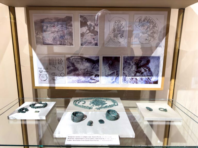 Wide view of a glass case containing green-blue ornaments, excavation photographs, and burial diagrams.