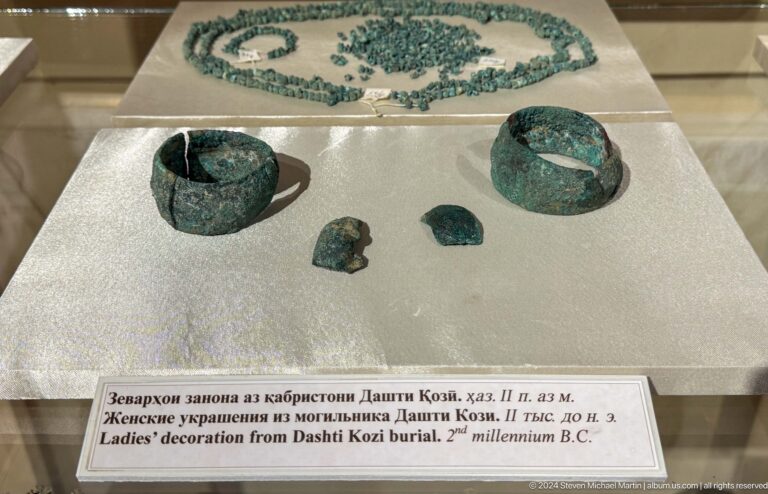 Two green-corroded bracelets, small ornaments, and a bead strand displayed above a bilingual label.