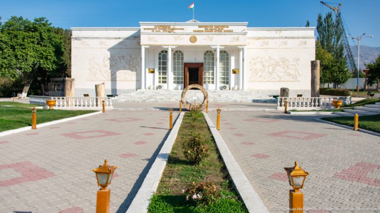 Central walkway and planted median leading to the white museum entrance beneath the Tajik flag.