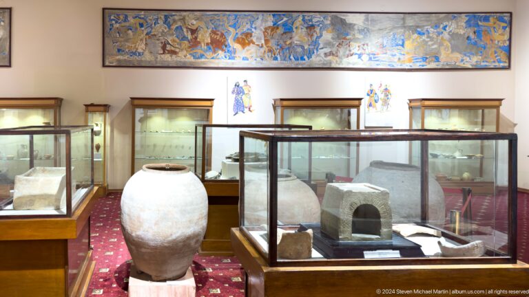 Wide museum gallery with glass cases, a large pale jar, stone objects, and a painted frieze.