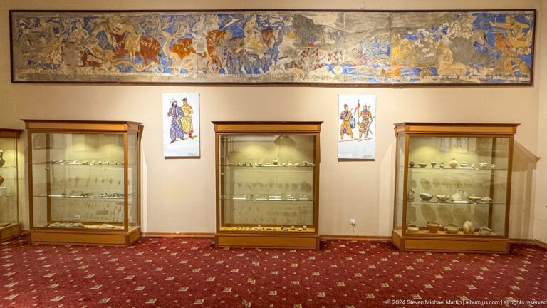 Symmetrical museum gallery with three pottery cases beneath a long ancient-style mural.