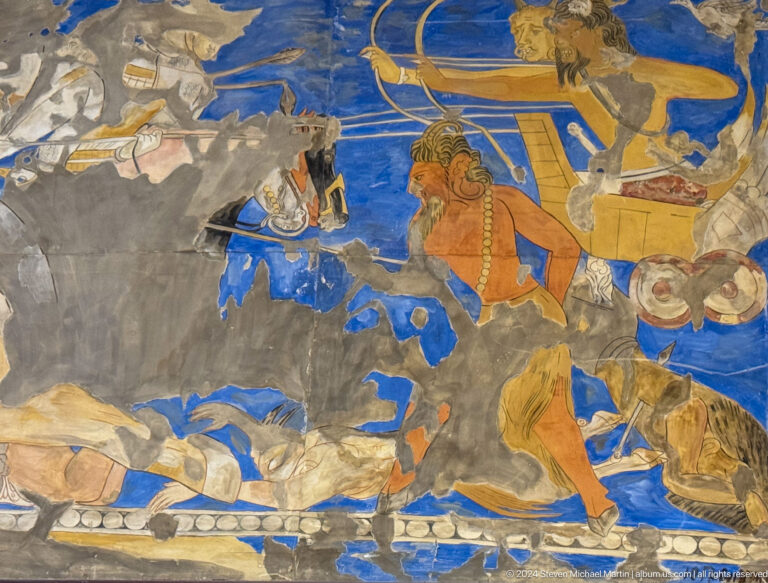 Fragmentary blue mural with large orange felines, weapons, and fallen human figures.