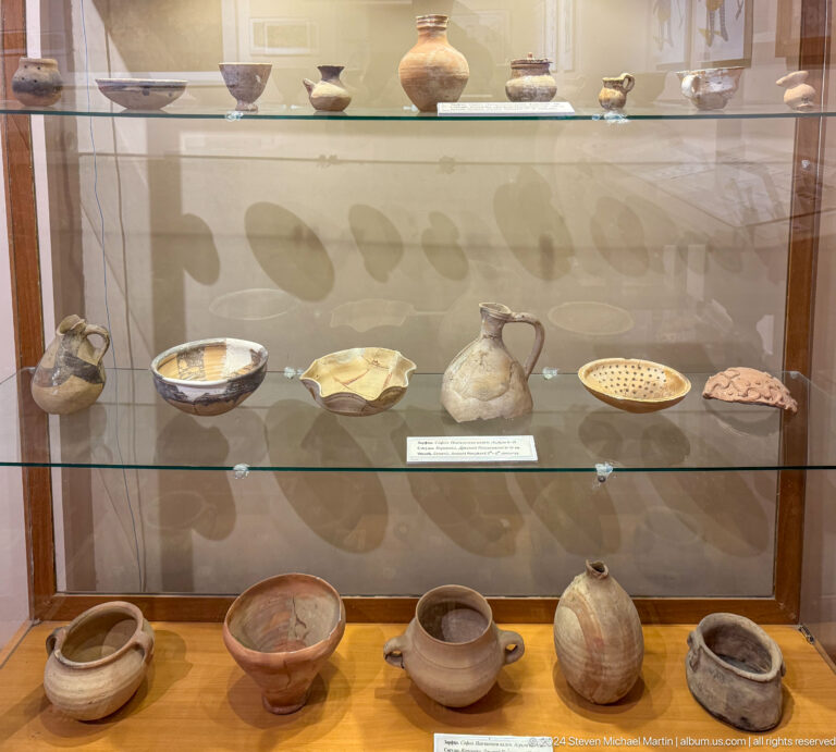 Glass case with numerous ceramic bowls, jars, lamps, and small vessels.