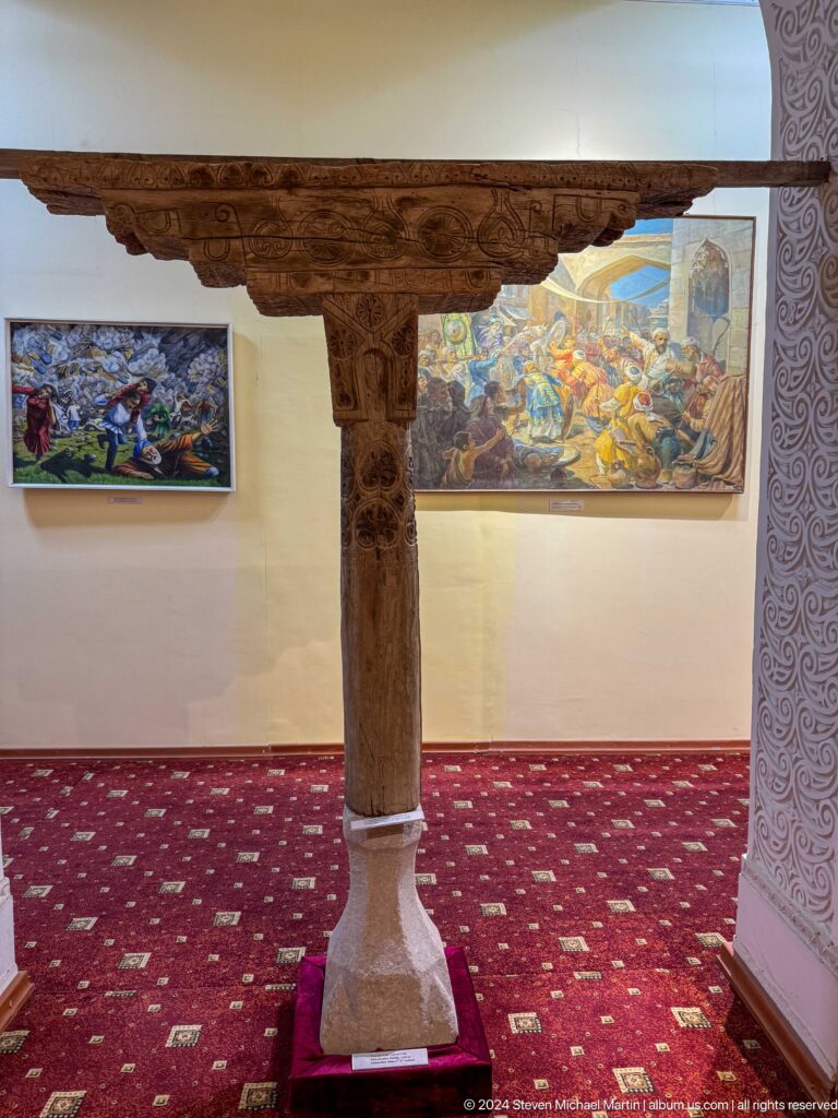 Full-length carved wooden column displayed between framed paintings in a museum gallery.