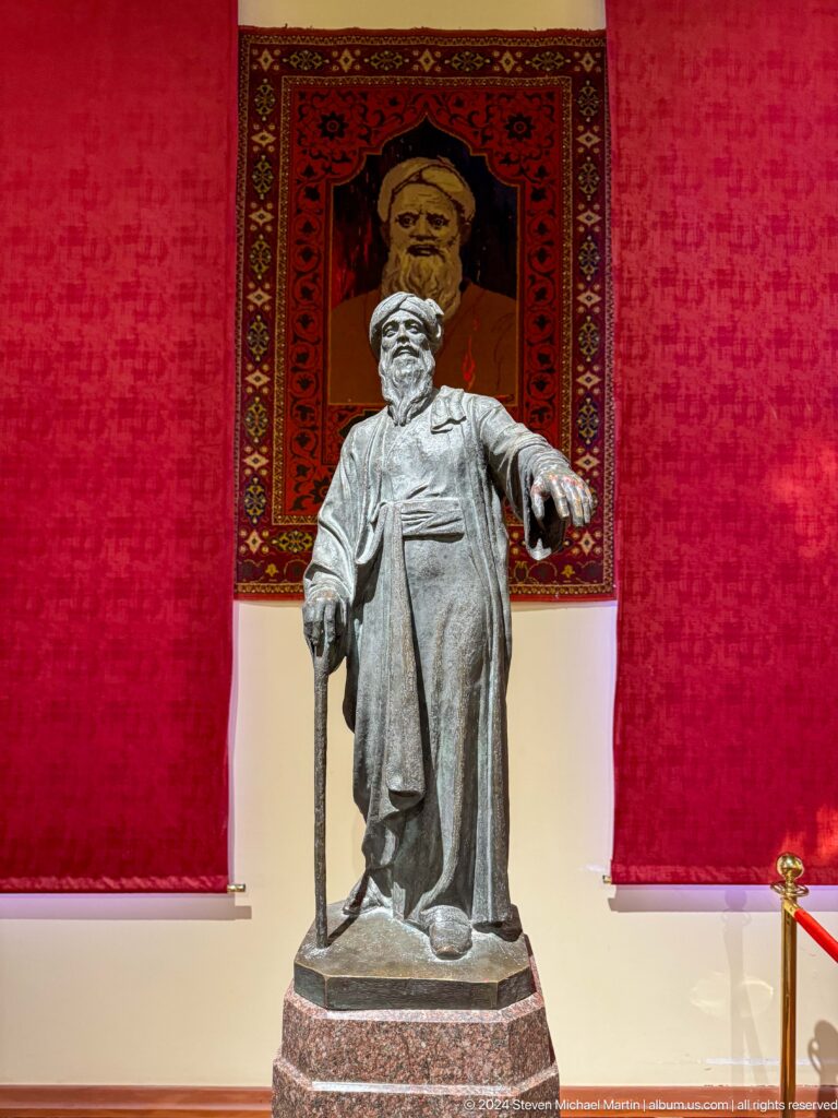 Gray standing statue of a robed man with one arm extended against a red textile.