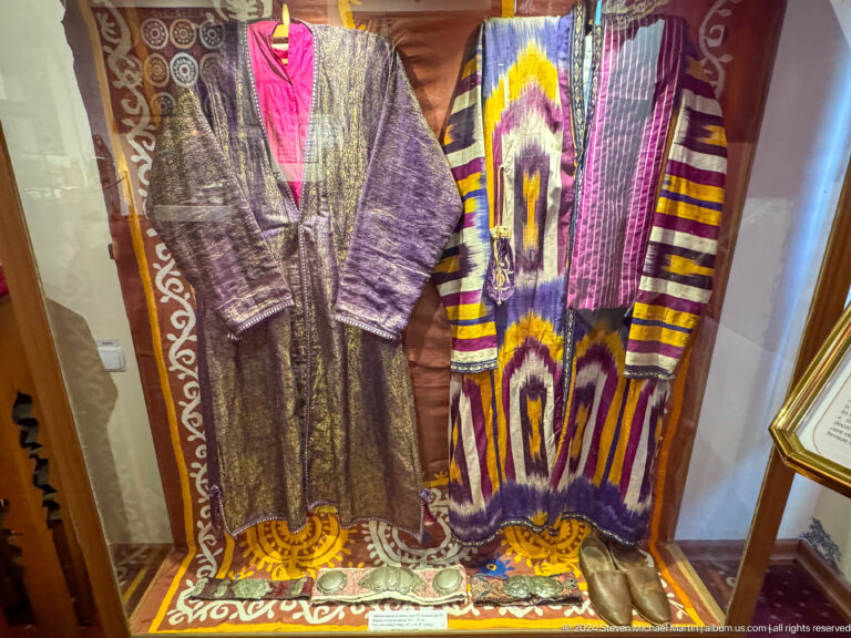 Long purple, gold, red, and white embroidered robes hanging inside a museum case.