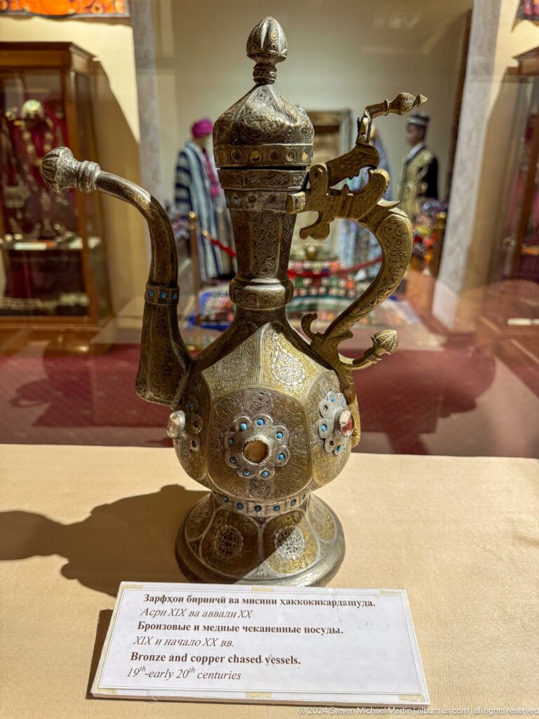 Close view of an engraved metal ewer with floral medallions and a curved handle.