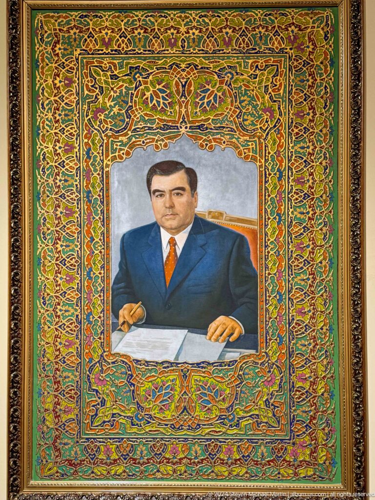 Formal portrait of a man in a dark suit and orange tie within an ornate frame.