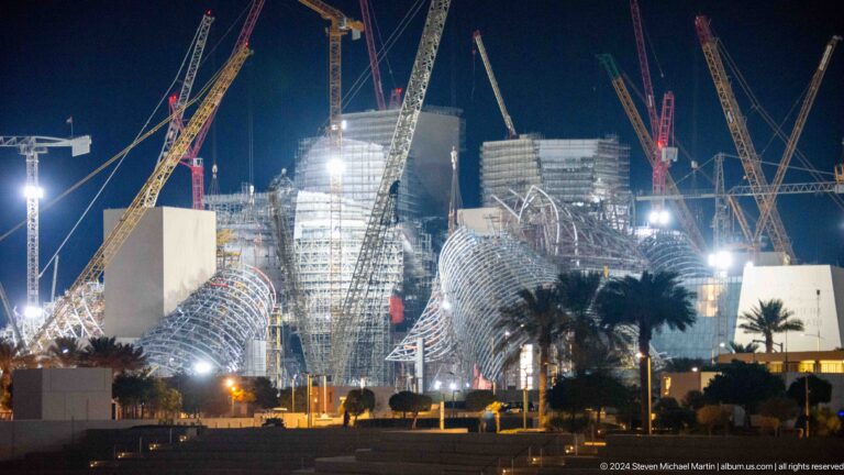Night view of a large, angular building under construction in Abu Dhabi’s Saadiyat Cultural District, illuminated floodlights revealing complex roof forms and multiple tower cranes.