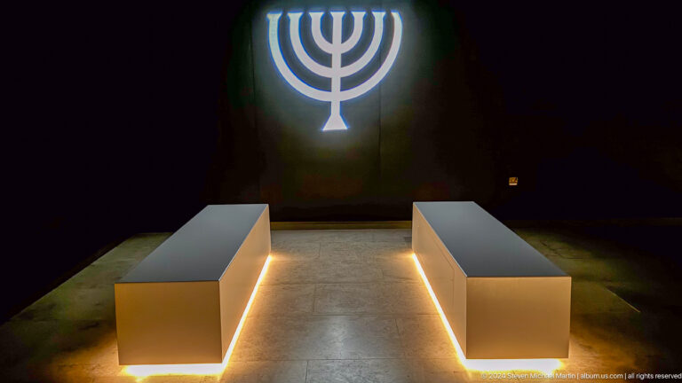 Dark reflection room with a large illuminated menorah on the back wall and two low benches outlined by warm floor lighting.