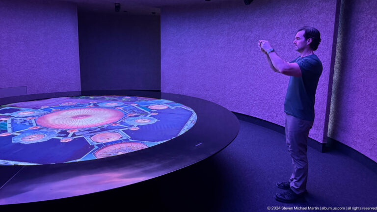 Man in a dim gallery reaching toward a large glowing circular digital projection on the wall at the Abrahamic Family House.