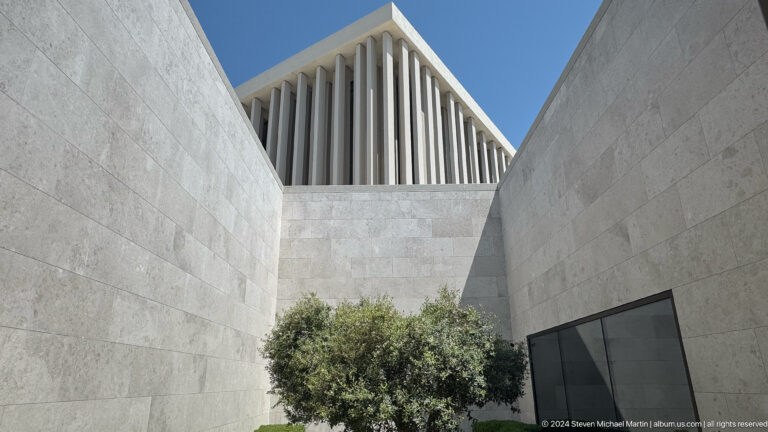 Daytime view of an olive tree centered in a recessed stone courtyard between pale walls at the Abrahamic Family House in Abu Dhabi, with vertical fins rising above the left wall.
