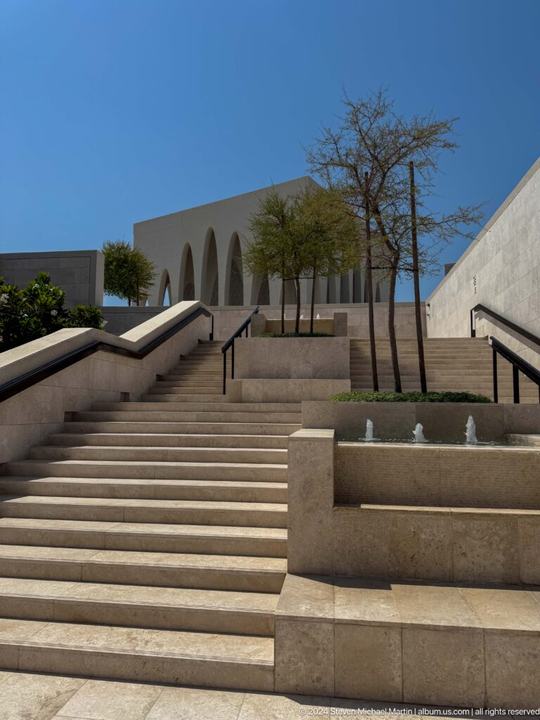 Daytime view looking up wide pale stone stairs at the Abrahamic Family House, flanked by accessible ramps, black handrails, and sparse trees against geometric buildings.