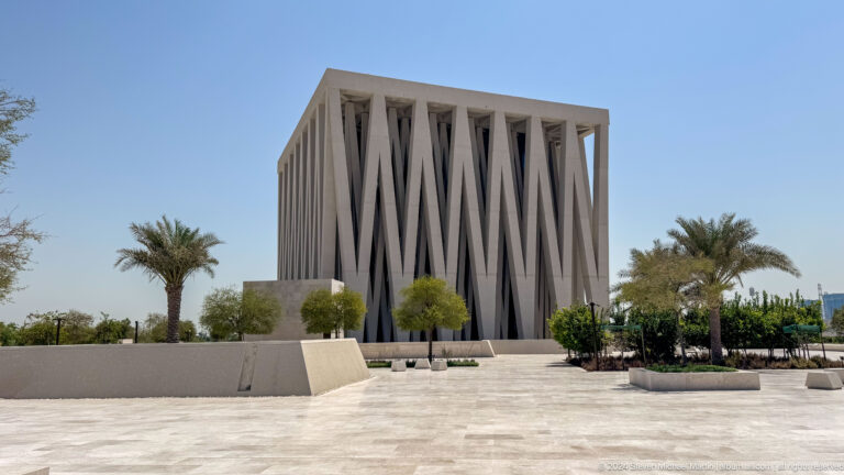 Daytime view of a cubic Abrahamic Family House worship building on Saadiyat Island, clad in diagonal facade fins and fronted by a wide paved plaza with palm trees in the foreground.