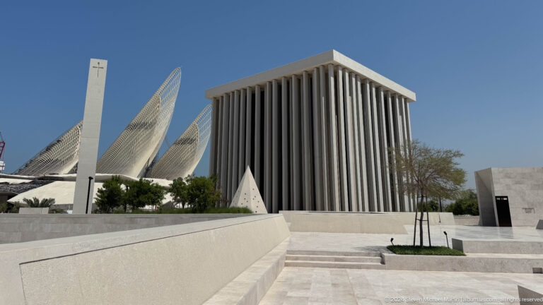 Wide view of the Abrahamic Family House in Abu Dhabi, showing a cubic worship building with deep vertical fins alongside tapering white forms rising beyond low courtyard walls.