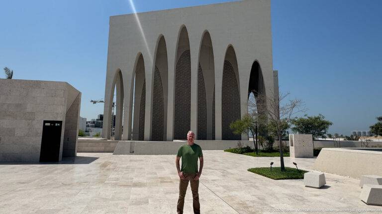Man in a green shirt standing in the paved forecourt before a row of very tall white pointed arches at the Abrahamic Family House in Abu Dhabi.