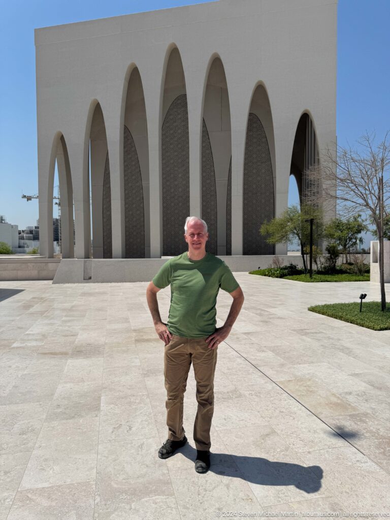 Man in a green T-shirt and light trousers stands with hands on hips before the large pointed arches of the Abrahamic Family House in Abu Dhabi.