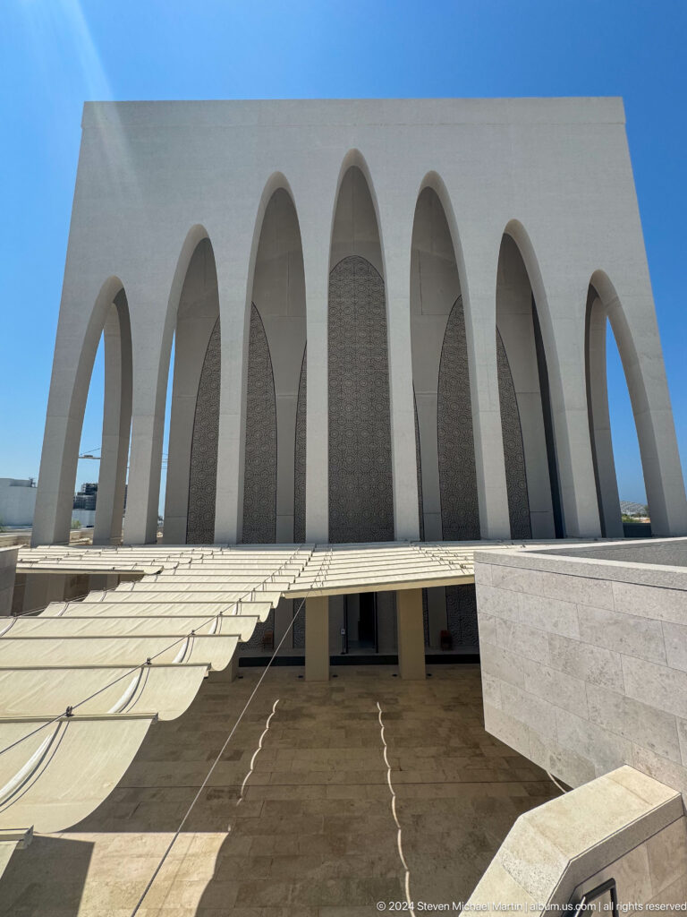Frontal upward view of the Abrahamic Family House worship building facade, showing multiple tall white pointed arches in deep shadow forming a rhythmic contemporary screen.