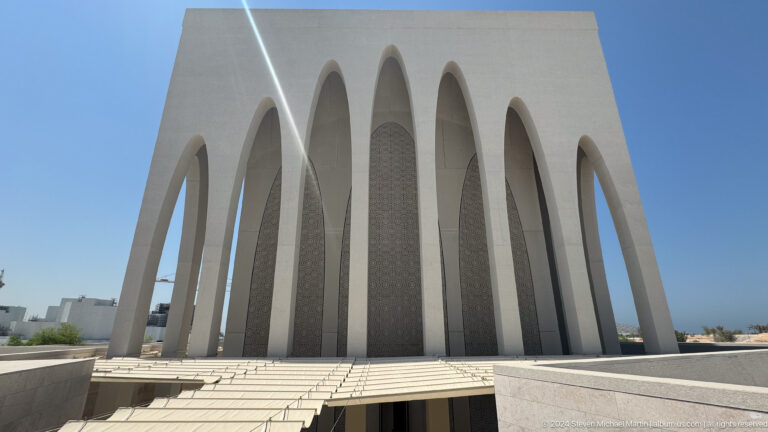 Frontal view of the Abrahamic Family House worship building exterior, showing an evenly spaced arcade of arches above steps leading to a shaded, recessed entrance.