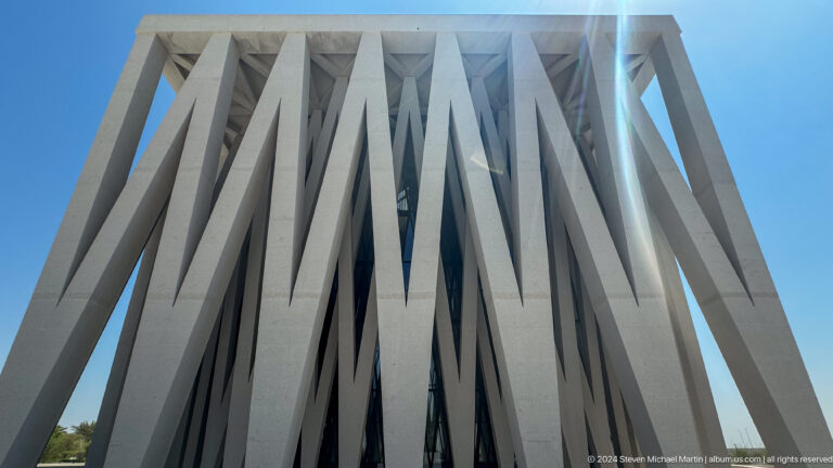 Low-angle view of overlapping white diagonal fins forming V-shapes across a dark glazed facade at the Abrahamic Family House.