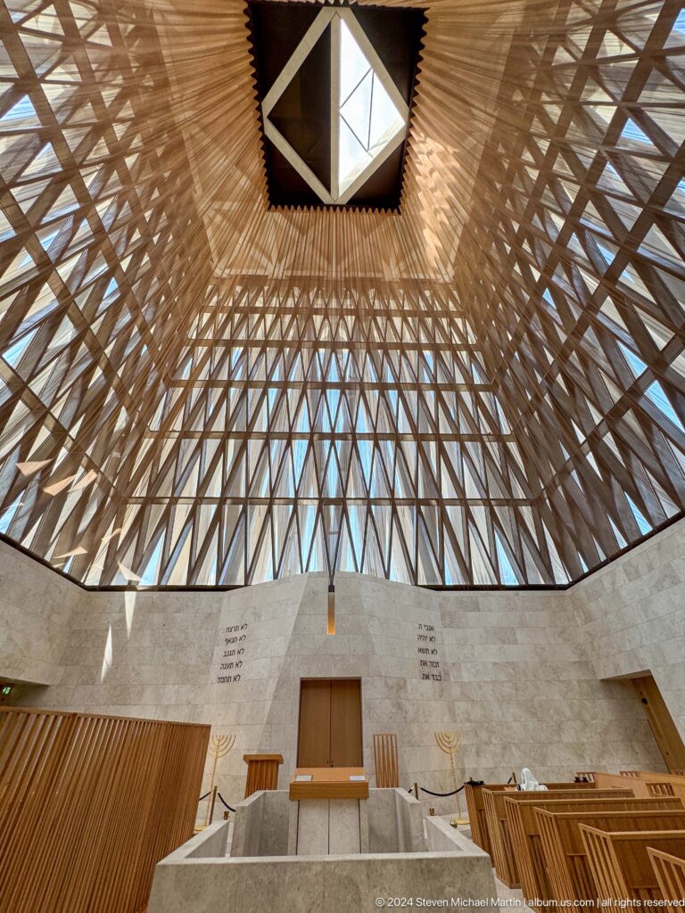 Upward view of the Abrahamic Family House worship interior ceiling, showing a warm-toned geometric lattice structure converging on a bright central opening.