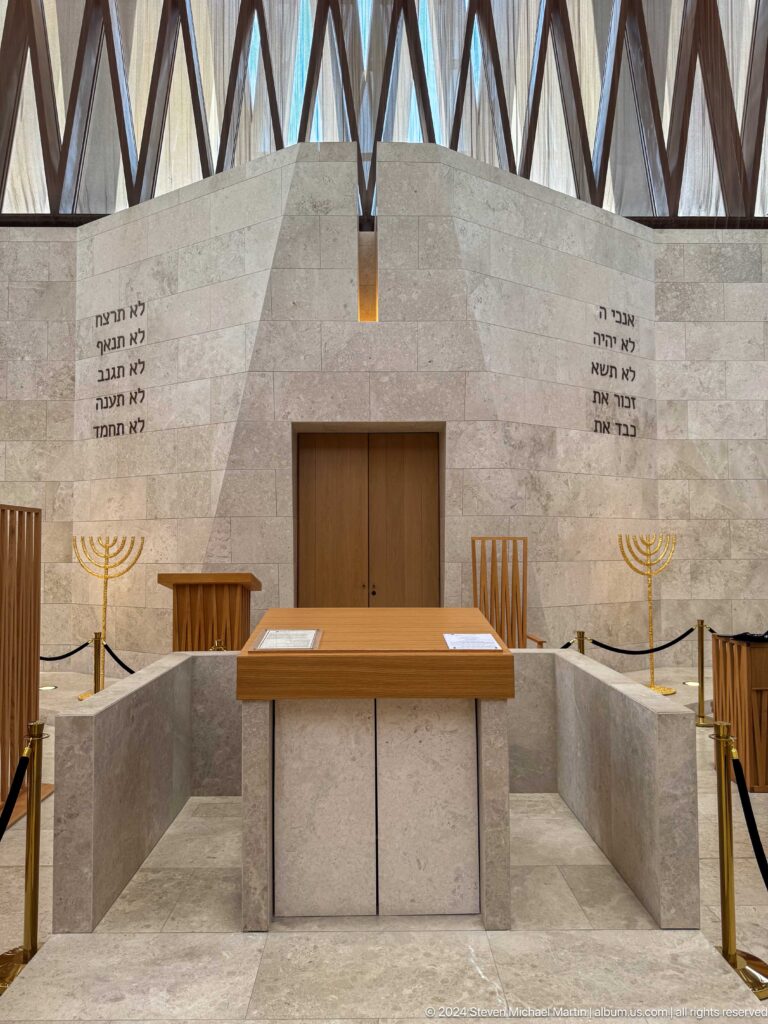 Interior of the Moses Ben Maimon Synagogue showing a central bimah with a wooden lectern, two menorahs, and Hebrew inscriptions carved into pale stone walls.