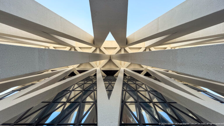 Upward view of the Abrahamic Family House roof structure with triangular white beams and dark glazing converging symmetrically toward a central point.