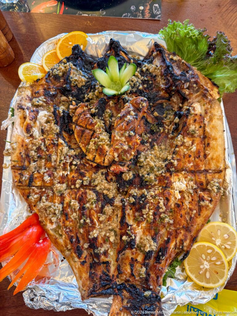 Overhead view of a butterflied whole grilled fish on a foil-lined metal tray, seasoned and charred with lemon wedges on the side.