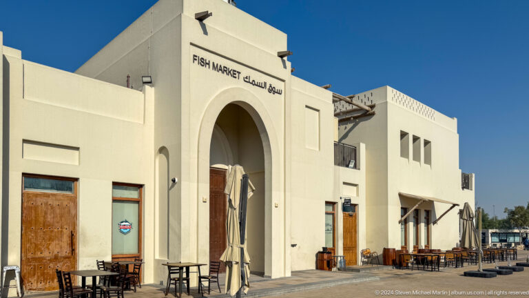 Daytime exterior of Al Dhafra Restaurant and Fish Market in Mina Zayed, Abu Dhabi, showing a low white building with a broad pointed entrance arch and bilingual signage near the roofline.