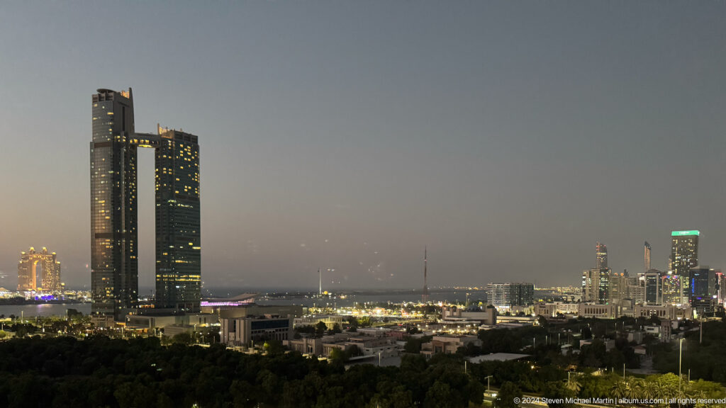 Telephoto view of Abu Dhabi at dusk, with illuminated mosque dome and minaret set amid high-rise towers and low urban buildings under a hazy sky.