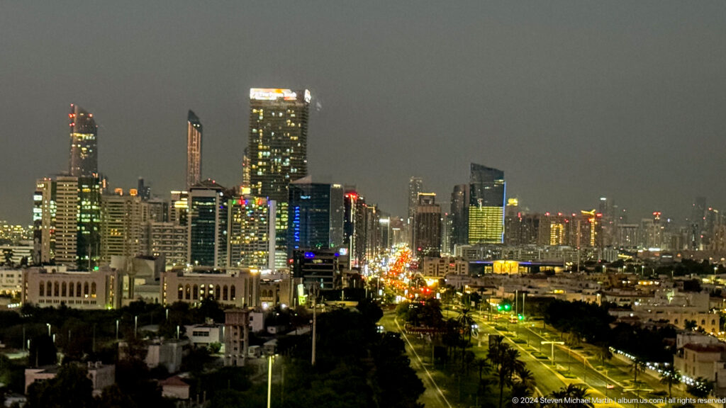 Elevated night view of central Abu Dhabi with illuminated high-rise towers and a brightly lit main road filled with traffic stretching into the distance.
