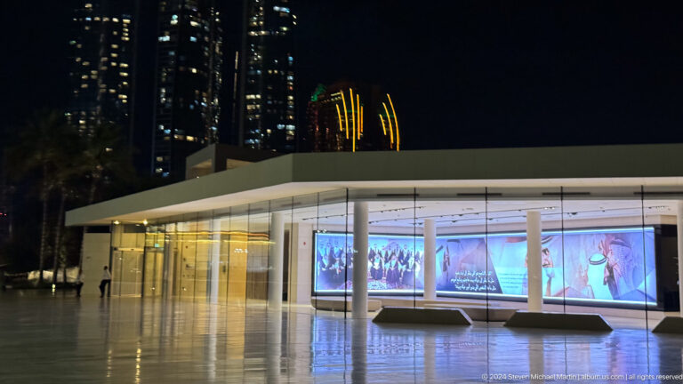 Night view of a low modern cultural pavilion in Abu Dhabi, with illuminated display screens visible through a glass façade along a dark, horizontal upper exterior.