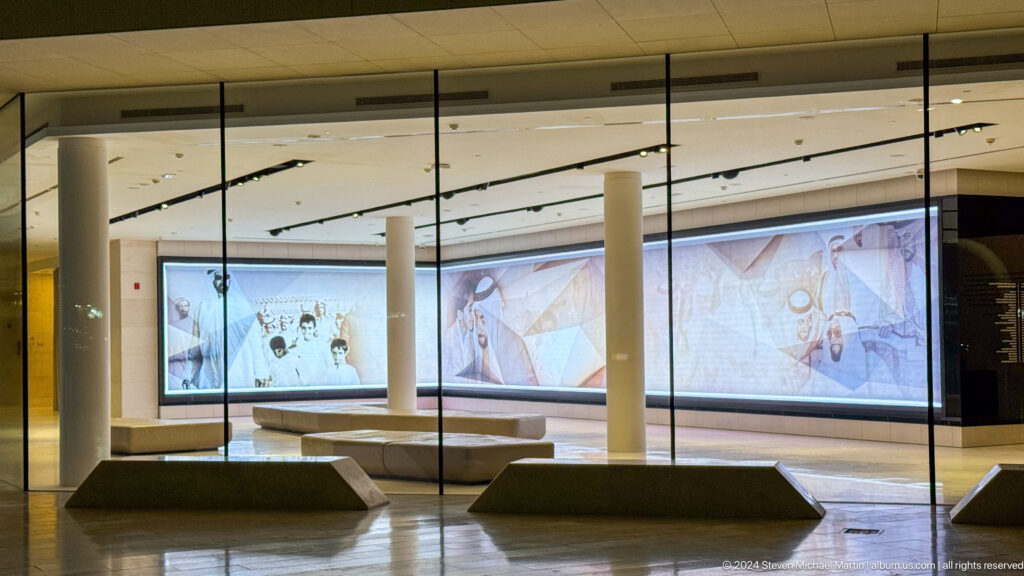Wide interior view of an immersive gallery in Abu Dhabi with a continuous row of bright digital screens along the back wall and a few dark angular benches on the floor.