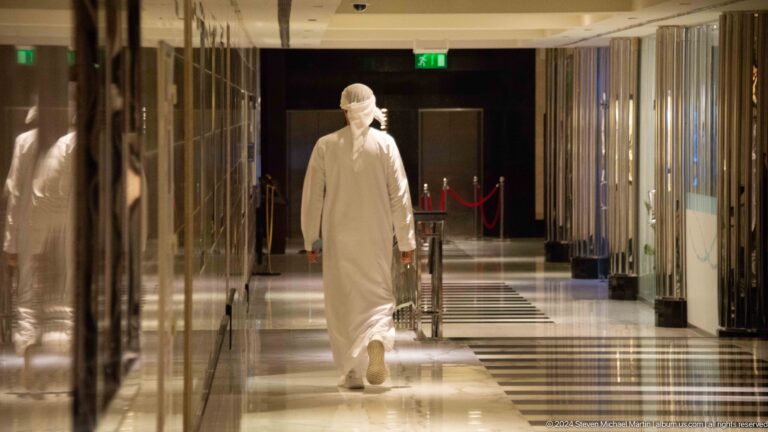 Emirati man in white kandura walking away along a brightly lit luxury hotel corridor with mirrored walls and polished floor.