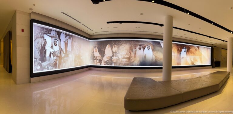 Curved gallery wall in a darkened room showing large-scale projected figures and landscapes, with a low bench following the arc of the screen.