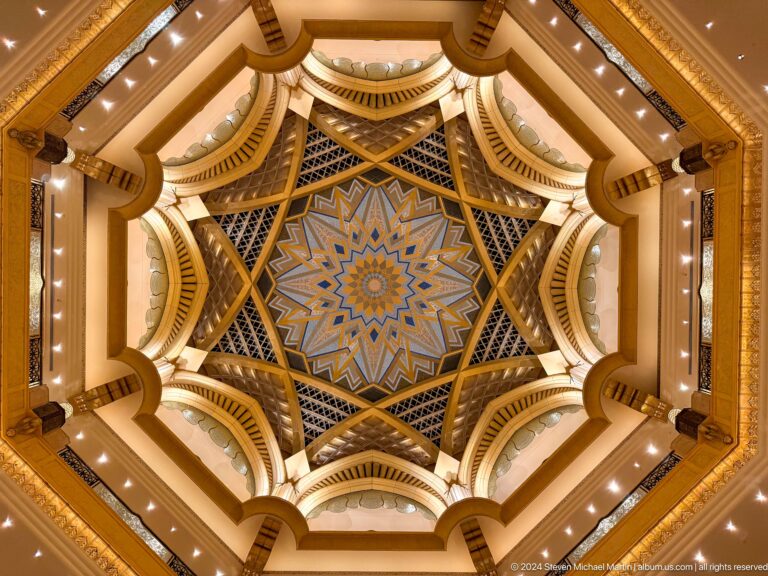 Centered upward view of the Emirates Palace gilded octagonal ceiling with a blue-and-gold star motif and concentric carved bands highlighted by warm lighting.