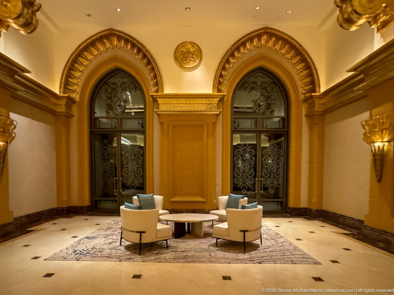 Symmetrical Emirates Palace lounge alcove with two pale armchairs and a small table set before tall arched windows and gold-paneled walls.