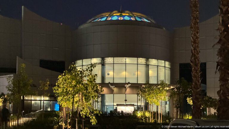 Night view of a circular building in Abu Dhabi with a glowing domed roof, dark exterior walls, glass entrance, and trees in the foreground.