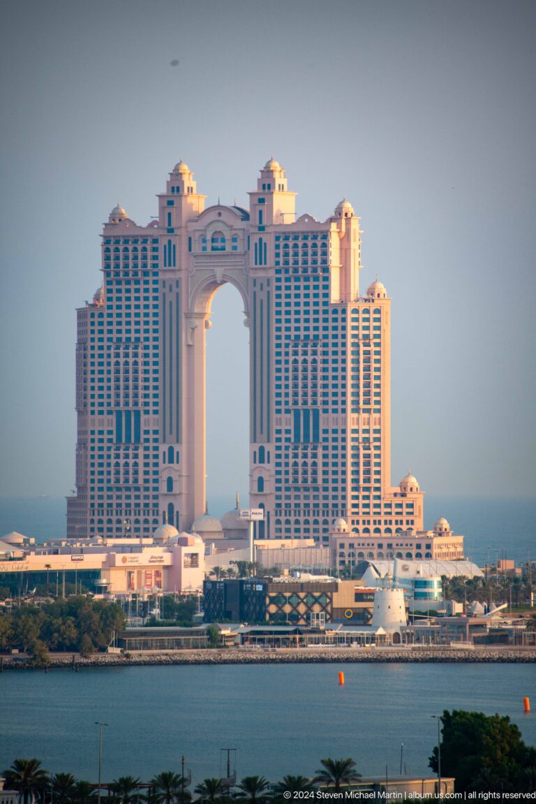 Daytime view of Fairmont Marina Abu Dhabi’s twin-tower hotel across the water, with a monumental open arch forming the central void between its pink and cream façades.