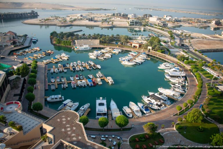 High daytime view of Abu Dhabi marina near Marina Mall, with numerous white yachts moored along curved docks in a sheltered turquoise basin bordered by landscaped roads.