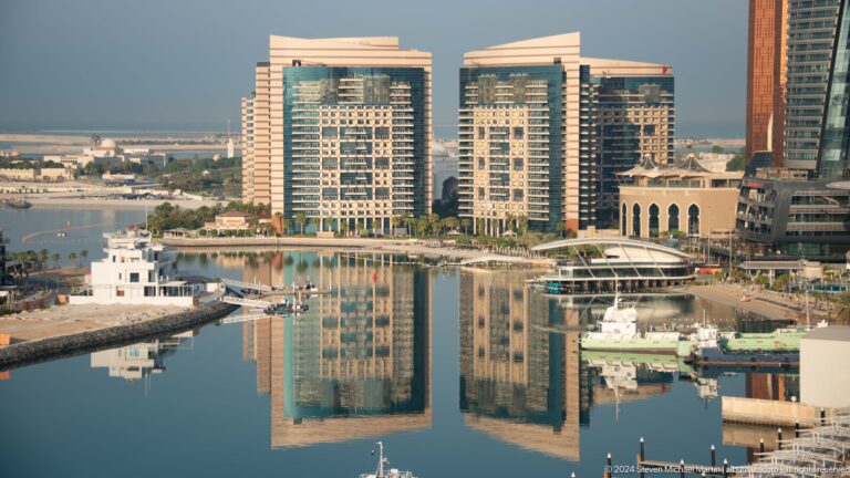 Telephoto view of three rectangular mid-rise waterfront buildings in Abu Dhabi reflected clearly in calm marina water.