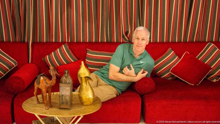 Man in a green shirt reclining on red striped cushions in a majlis-style seating area beside brass dallah coffee pots and other decorative objects.