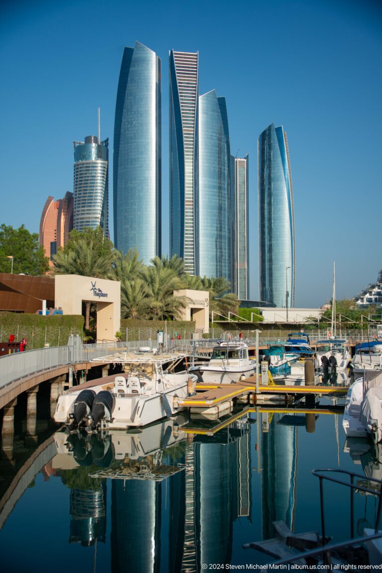 Daytime view of small boats moored at an Abu Dhabi marina with landscaped promenades in the foreground and the Etihad Towers rising behind.