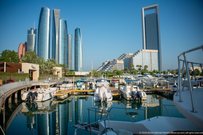 Wide daytime view across calm water at an Abu Dhabi marina, where white yachts and waterfront high-rises are reflected clearly in the still surface.