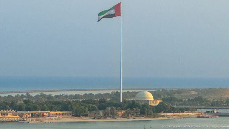 Telephoto view of a very large UAE flag flying above a low green shoreline with a distant golden dome on the horizon in Abu Dhabi.
