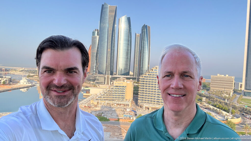 Two men smile at the camera in a close selfie at the Abu Dhabi waterfront, with the Etihad Towers visible in the background.
