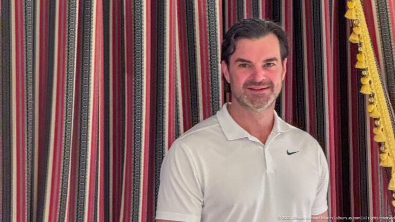Waist-up portrait of a smiling man in a white polo shirt standing in front of traditional red-and-black striped majlis fabric.