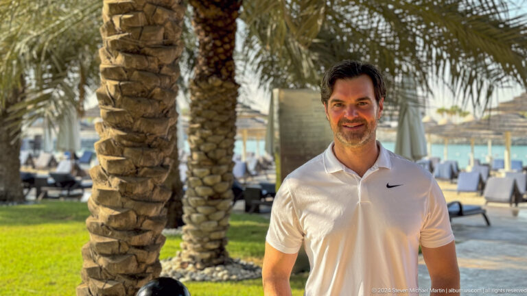 Smiling man in a white polo shirt standing by a palm tree in a landscaped resort area with green lawn in Abu Dhabi.