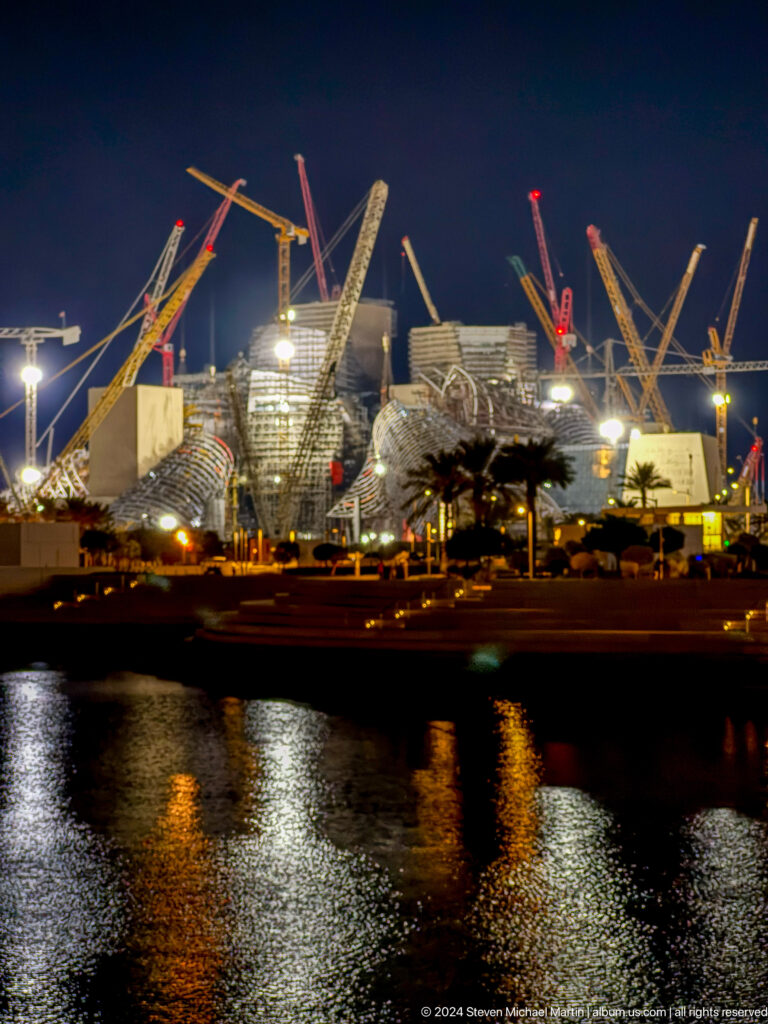 Nighttime view of Saadiyat Cultural District construction site in Abu Dhabi with cranes and illuminated faceted buildings reflected in the dark waterfront.