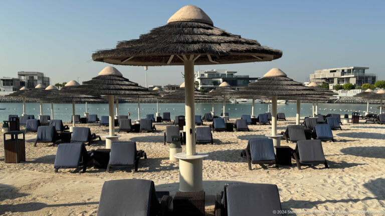 Empty dark loungers under large woven parasols on a sandy Abu Dhabi beach beside calm blue water.